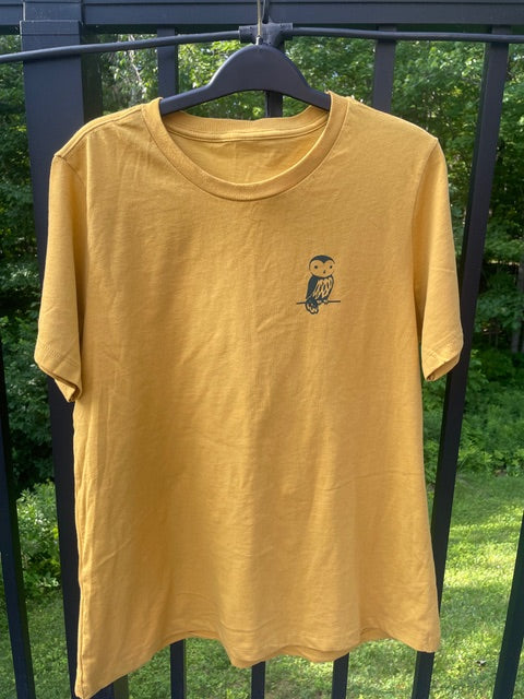 Yellow t-shirt with Barred Woods logo on a hanger against a green outdoor background.