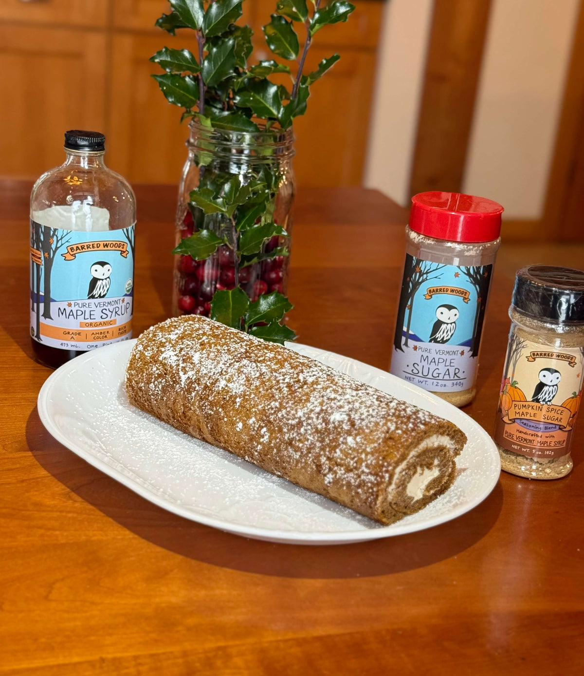 A maple roll cake on a plate with jars of maple syrup, maple sugar, and Barred Woods Pumpkin Spice .